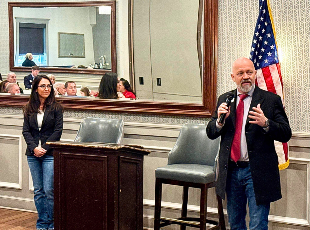 Senator Scott Bright speaking at a Brighton, Colorado community event addressing District 13 constituents with an American flag in the background.